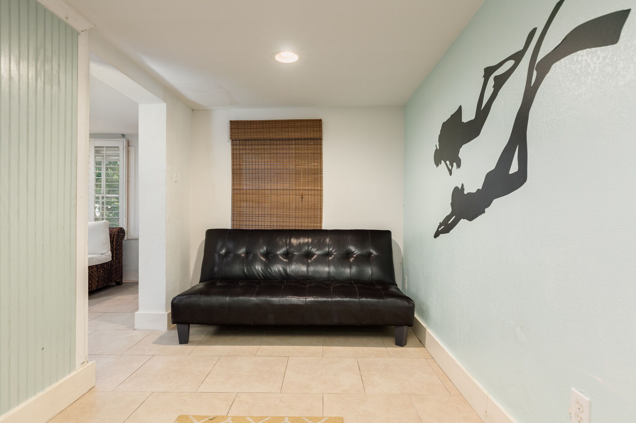 Une pièce avec un canapé en cuir noir, un mur orné d’une fresque représentant une silhouette noire, une fenêtre avec des stores et une porte donnant sur une autre pièce.