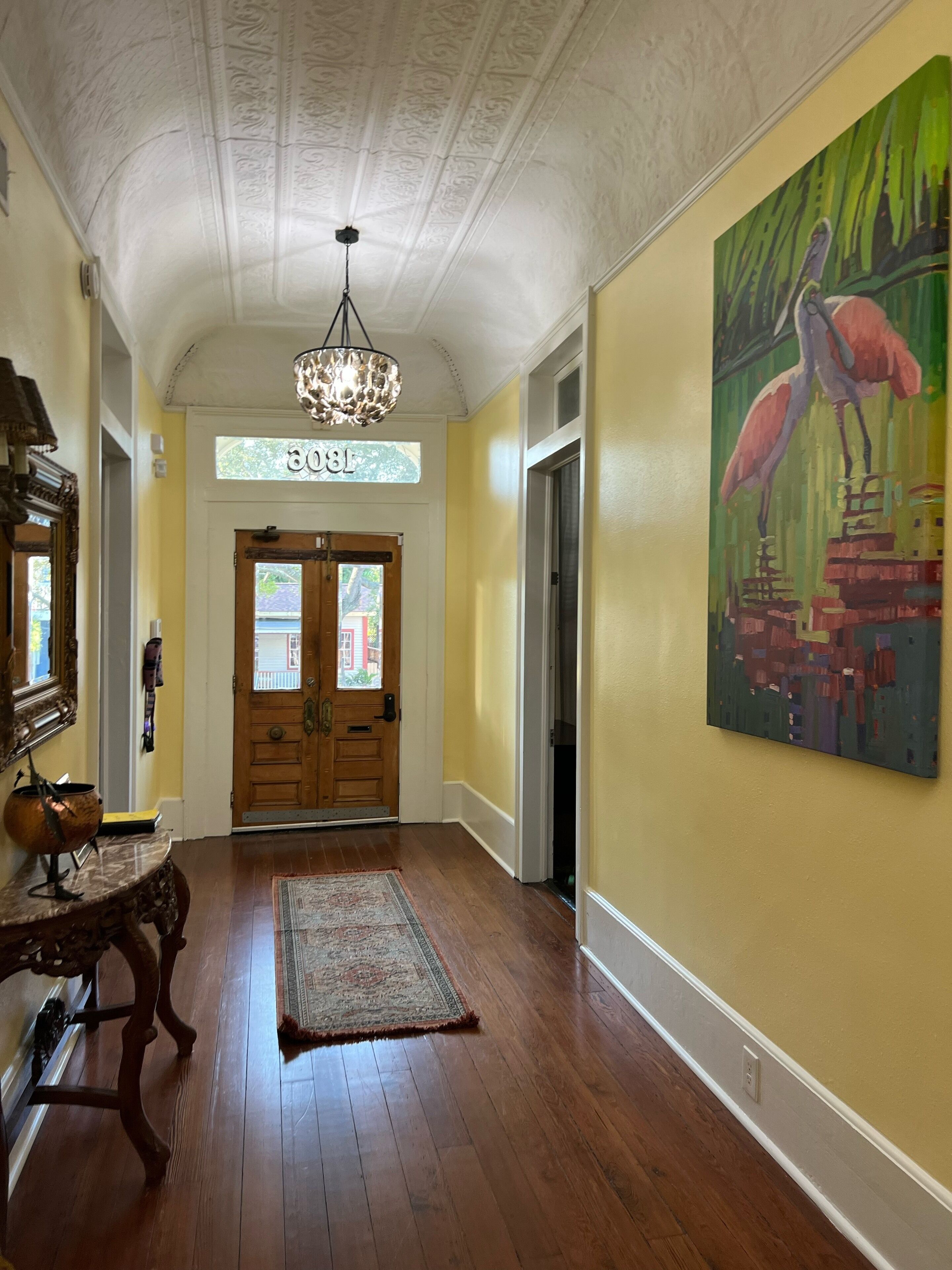 A hallway with wooden floors, a chandelier, a painting of flamingos, and a door with a number sign.