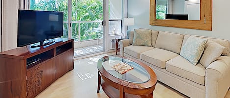 Ein modernes Wohnzimmer mit einem Flachbildfernseher, einem Holzschrank, einem Glastisch, einem beigen Sofa und einem großen Fenster mit Blick auf Grünflächen.