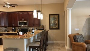 A kitchen with dark wooden cabinets, a stainless steel microwave, and a granite countertop. There is a dining area with a wicker chair and a sofa with leopard print cushions. - Newer Upscale Home with Htd Pool & Spa - Bike or Walk to Beaches and Restaurants (Naples)