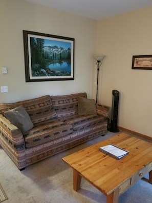 A living room with a patterned sofa, a wooden coffee table, a framed landscape painting, a floor lamp, and a small table with a document. - Great Location, Heated Slate Floors (Whistler)