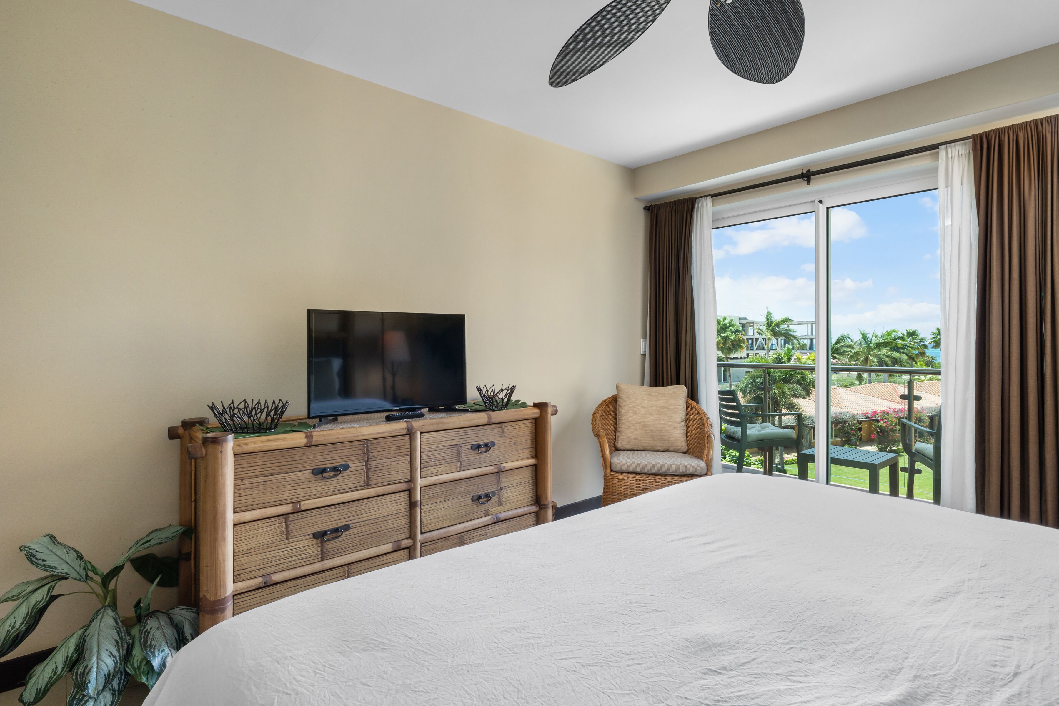 A hotel room with a large bed, a flat-screen TV on a wooden dresser, a wicker chair, and a balcony with a view of palm trees and a building.