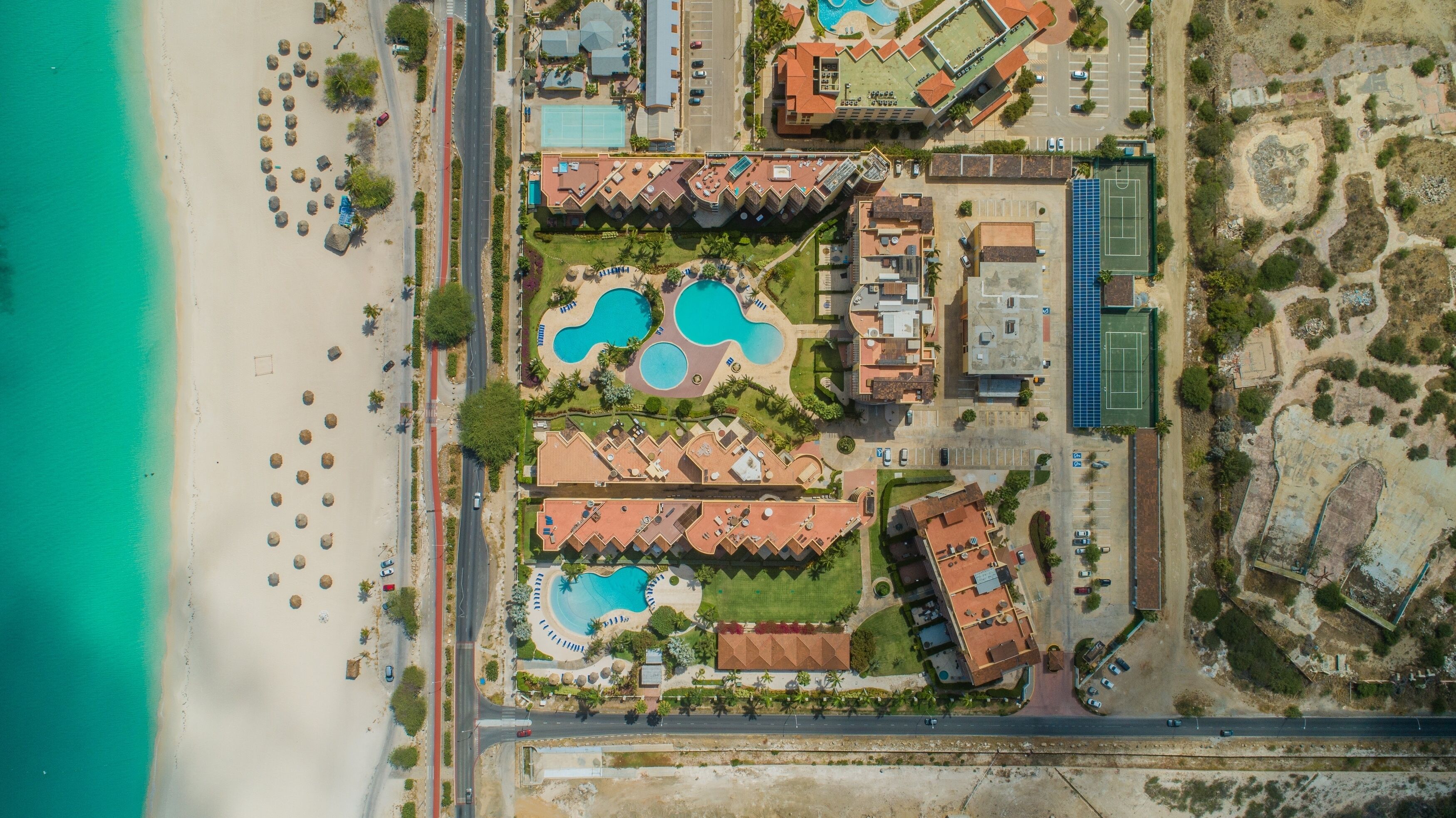 An aerial view of a resort with multiple swimming pools, beachfront, and a road running through the property.