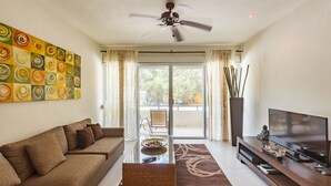 A modern living room with a sofa, a glass coffee table, a TV, and a ceiling fan. - Meridian Beach Condo, 200m from beach and 5th avenue, Wifi (Playa Del Carmen)