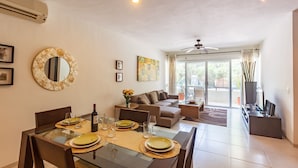 A dining area with a glass table set for four, a sofa, and a large window with a view of greenery. - Meridian Beach Condo, 200m from beach and 5th avenue, Wifi (Playa Del Carmen)