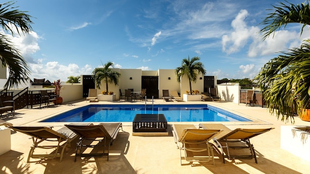 A swimming pool with lounge chairs and palm trees, surrounded by a white building with a flat roof. - Meridian Beach Condo, 200m from beach and 5th avenue, Wifi (Playa Del Carmen)