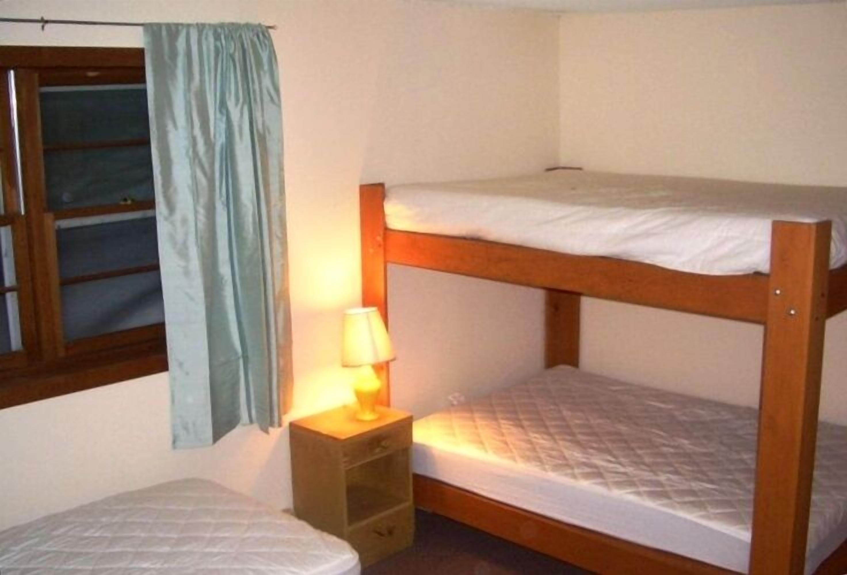 A bunk bed with a bedside table and lamp, a window with curtains, and a small bedside table with drawers.