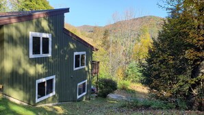A green wooden cabin with three windows and a deck, surrounded by autumn foliage and hills. - Killington area home - 4-seasons, hot tub and A/C (Pittsfield)