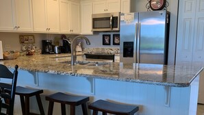 Eine Küche mit weißen Schränken, einem Edelstahl-Kühlschrank, einer Mikrowelle und einer Kaffeemaschine. An der Theke stehen drei Holzstühle.