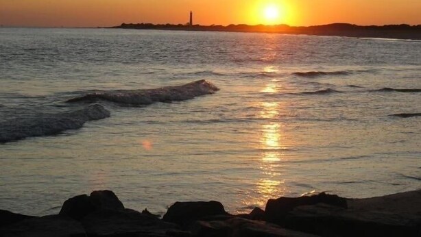 A sunset over a calm sea with waves, a distant lighthouse, and rocky shoreline.