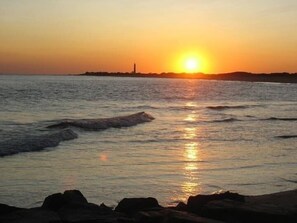 A sunset over a calm sea with waves, a distant lighthouse, and rocky shoreline. - Cozy Condo. 3 BR, 2 BA. Close to Beach and Shopping. Reserved parking spot. (Cape May)