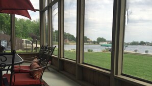 Une pièce dotée de grandes fenêtres donnant sur un lac, un espace patio avec une table et des chaises, et une vue sur une zone herbeuse et des bâtiments situés au loin.
