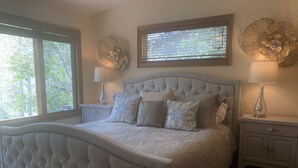 Een slaapkamer met een gestoffeerd hoofdeinde, een groot raam met zonwering, een nachtkastje met een lamp en een decoratief wandobject.