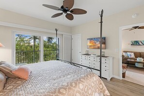 Ein Schlafzimmer mit Bett, Deckenventilator, Kommode, Gemälde und einem Balkon mit Blick auf Bäume.