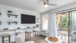 Un salon moderne doté d’un téléviseur à écran plat, de tabourets de bar en bois, d’un canapé blanc et d’un balcon offrant une vue sur l’extérieur.