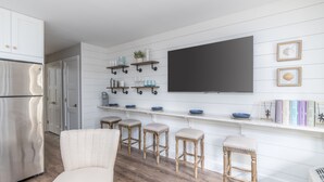 Une cuisine moderne avec des meubles blancs, un grand téléviseur à écran plat, des tabourets en bois et un coin bar doté d’étagères.