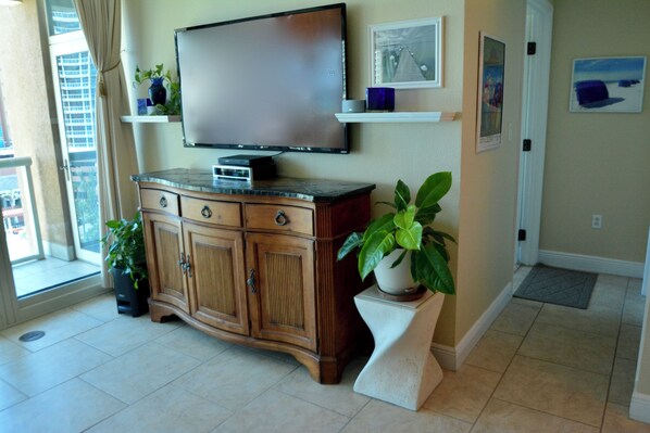 Een woonkamer met een flatscreen-televisie aan de muur, houten kasten, een potplant en een deur naar een balkon.