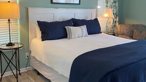 A bedroom with a bed, a headboard, a nightstand, a lamp, a window with blinds, a ceiling fan, and a framed picture on the wall.
