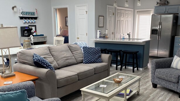 A modern living room with a sofa, coffee table, and patterned chairs. A kitchen area with a refrigerator and bar stools is visible in the background.