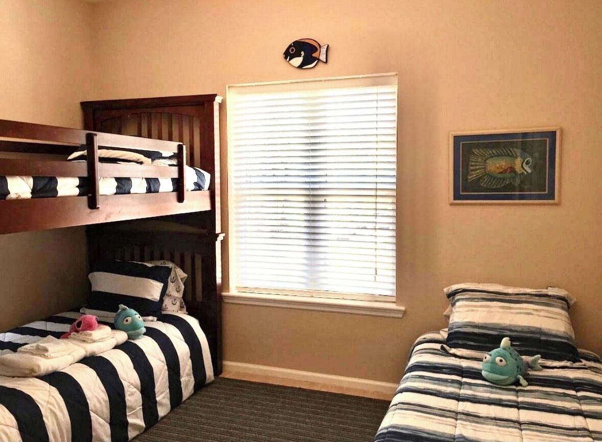 Ein Schlafzimmer mit zwei Betten, davon ein Etagenbett und ein Einzelbett. Es gibt ein Fenster mit Jalousien, eine Fischdekoration an der Wand und ein gerahmtes Bild.