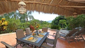 An outdoor dining area with a table set for four, surrounded by chairs, under a thatched roof.