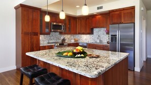 A kitchen with wooden cabinets, a granite countertop, a stainless steel refrigerator, and a central island with stools. - Pili Mai 11I: Tropical Condo w/ Fairway Views, AC, Shared Pool & Jacuzzi (Koloa)