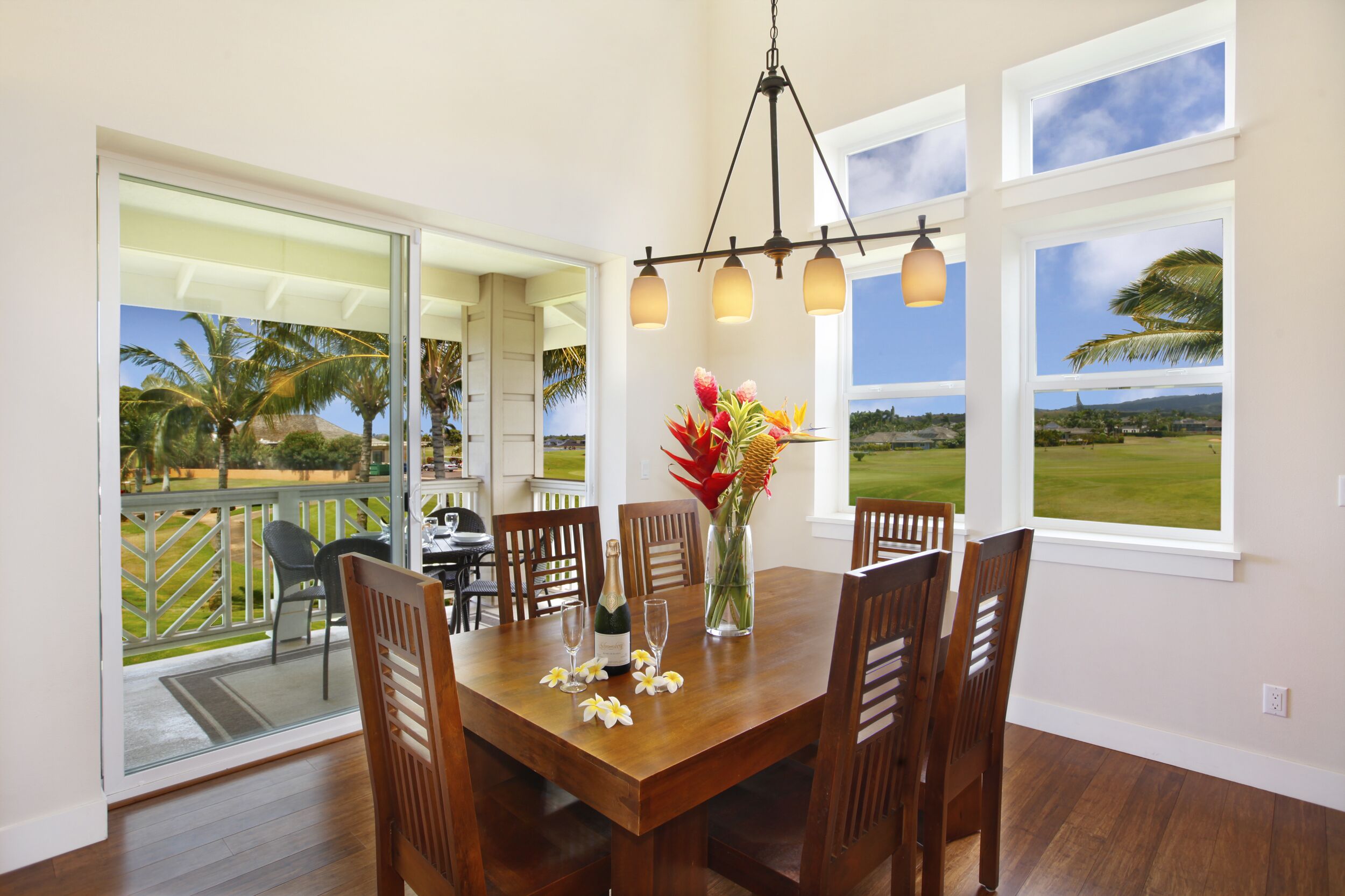 A dining area with a wooden table, chairs, a chandelier, and a view of a tropical landscape through large windows.