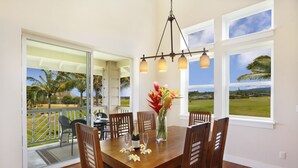 A dining area with a wooden table, chairs, a chandelier, and a view of a tropical landscape through large windows. - Pili Mai 11I: Tropical Condo w/ Fairway Views, AC, Shared Pool & Jacuzzi (Koloa)