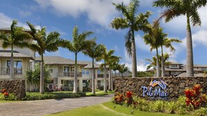 A hotel entrance with a stone wall, palm trees, and a sign that reads "Pili Mai." - Pili Mai 11I: Tropical Condo w/ Fairway Views, AC, Shared Pool & Jacuzzi (Koloa)