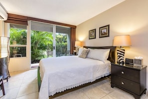 A bedroom with a large bed, two bedside lamps, a nightstand, and a view of greenery outside.