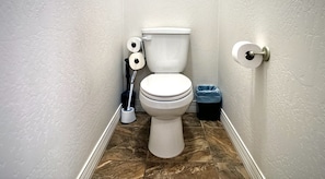 A bathroom with a toilet, two rolls of toilet paper, a plunger, and a trash bin.