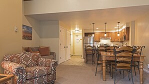 A living room with patterned furniture, a dining area with wooden chairs and a table, and a kitchen with wooden cabinets and a refrigerator.