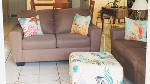 A living room with a sofa, ottoman, and chairs. There is a dining table with chairs in the background. - GATED OCEANFRONT CONDO AT SMATHER'S BEACH (Key West)