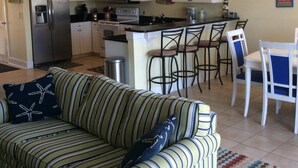 Eine moderne Küche mit weißen Schränken, einem Edelstahl-Kühlschrank, einer Mikrowelle und einem Herd. Es gibt einen Essbereich mit einem weißen Tisch und Stühlen, sowie ein gestreiftes Sofa mit Kissen.