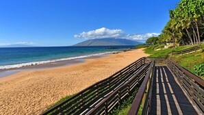 Une plage avec une promenade en bois qui mène au bord de l’eau, un ciel bleu dégagé et une montagne à l’horizon.