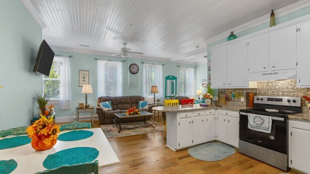 Una sala de estar acogedora con sofá, mesa de centro y televisor. La cocina cuenta con armarios blancos, estufa y un comedor con mesa y sillas.