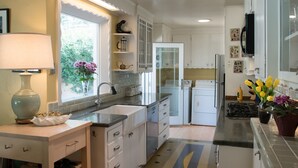 Eine moderne Küche mit weißen Schränken, einer Edelstahl-Spüle und einem eingebauten Backofen. Ein Fenster bietet Blick auf grüne Vegetation, und auf der Arbeitsplatte steht eine Vase mit Blumen.