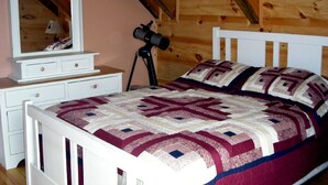 Een slaapkamer met een houten plafond, een wit bed met een geborduurde quilt, een dressoir met spiegel en een telescoop op een statief.