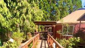 Un puente de madera con barandillas que conduce a un espacio cubierto con vistas a árboles y un edificio al fondo.