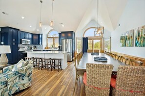 A modern kitchen with dark blue cabinets, a central island, and a dining area with wicker chairs. - Free Pool Heat on All New 2026 Reservations (Corolla)