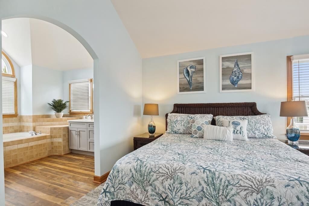 A bedroom with a large bed, two bedside lamps, a nightstand, a window with blinds, and a painting on the wall.