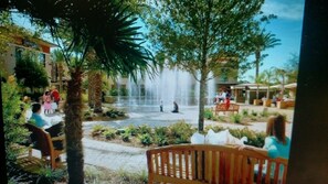 Relax in a serene oasis surrounded by lush greenery and palm trees, with a soothing fountain and comfortable benches.
