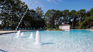 Una piscina con chorros de agua, rodeada de árboles y un edificio al fondo.