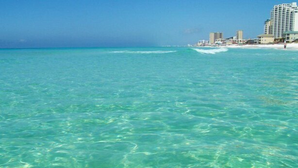 Una playa con vista despejada, agua turquesa y un hotel al fondo.