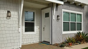 Ein modernes Haus mit weißer Tür, einem Fenster mit grünen Fensterläden und einem kleinen Garten mit Pflanzen und Steinen.