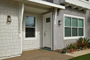 Ein modernes Haus mit weißer Tür, einem Fenster mit grünen Fensterläden und einem kleinen Garten mit Pflanzen und Steinen.