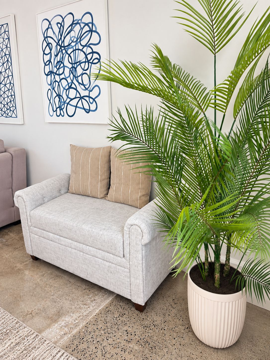 A light grey sofa with two pillows, a large potted plant with green fronds, and abstract art on the wall.