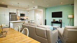Una cucina moderna con mobili bianchi, un frigorifero in acciaio inox e un forno a microonde. È presente una zona pranzo con tavolo e sedie in legno, e un soggiorno con un divano beige e una televisione a parete.
