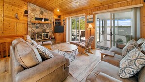 A cozy cabin interior with a stone fireplace, leather sofas, a television, and a view of a lake through large sliding glass doors. - Executive Lake Home on Premier Lake w Game Room & Sauna–Ultimate Retreat Awaits! (Webster)