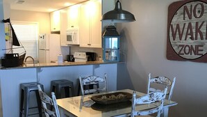 A compact kitchen with white cabinets, a dining table with white chairs, and a "NO WAKE ZONE" sign on the wall. - Updated, spacious Gulf-front Condo- just steps to the beach! (Panama City Beach)